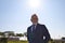 Handsome mature man, executive, grey haired, with beard, jacket and tie, is in a park posing for photo with sun in background.