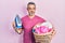 Handsome middle age man with grey hair holding electric steam iron holding laundry basket puffing cheeks with funny face