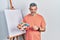 Handsome middle age man with grey hair standing drawing with palette by painter easel stand skeptic and nervous, frowning upset
