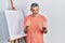 Handsome middle age man with grey hair standing by painter easel stand doing money gesture with hands, asking for salary payment,