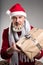 Handsome middle aged man with decorated beard wearing Christmas hat looking at camera, outstretched hands with gift box