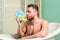 Handsome muscular man relaxing bathtub with yellow duck. Macho with sponge take bath at home. Taking bath with soap suds