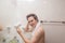 Handsome naked young man drying hair with hairdryer, looking at mirror at home