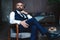 Handsome pensive man is touching his beard, looking away and thinking while sitting in armchair indoors.