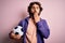 Handsome player man with beard playing soccer holding football ball over pink background cover mouth with hand shocked with shame