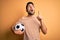 Handsome player man with beard playing soccer holding footballl ball over yellow background amazed and surprised looking up and