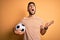 Handsome player man with beard playing soccer holding footballl ball over yellow background crazy and mad shouting and yelling