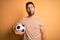 Handsome player man with beard playing soccer holding footballl ball over yellow background making fish face with lips, crazy and