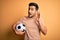 Handsome player man with beard playing soccer holding footballl ball over yellow background smiling with hand over ear listening