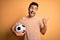 Handsome player man with beard playing soccer holding footballl ball over yellow background smiling with happy face looking and