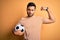 Handsome player man with beard playing soccer holding footballl ball over yellow background Strong person showing arm muscle,
