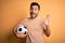 Handsome player man with beard playing soccer holding footballl ball over yellow background Waiving saying hello happy and