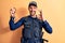 Handsome policeman wearing uniform and bulletprof holding handcuffs over yellow background doing ok sign with fingers, smiling