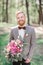 Handsome red bearded groom stands with pink bouquet in the wood
