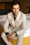 Handsome respectable groom is sitting on the bed of a luxury hotel room, the groom\'s light wedding suit