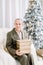 Handsome retired man in suit and shirt, holding present box, while sitting in white armchair in front of beautiful