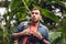 Handsome scared traveler with binoculars and backpack in green tropical forest.