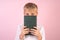 Handsome schoolboy with blonde hair covers his face with book while standing in front of pink background