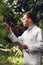 Handsome scientist in white coat and goggles examining plants in green orangery.