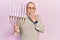 Handsome senior man with beard holding menorah hanukkah jewish candle covering mouth with hand, shocked and afraid for mistake