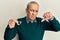 Handsome senior man with grey hair holding medical hearing aid with angry face, negative sign showing dislike with thumbs down,