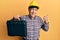 Handsome senior man with grey hair wearing safety helmet holding toolbox pointing thumb up to the side smiling happy with open