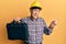 Handsome senior man with grey hair wearing safety helmet holding toolbox smiling happy pointing with hand and finger to the side