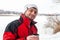 Handsome smiling bearded man in a red and black jacket and white cap holding a metal cup with hot tea against frozen river