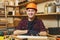 Handsome smiling young man working in carpentry workshop at wooden table place with piece of wood