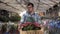 Handsome, tall male gardener in shirt and green apron carrying carton box with pink flowers plants while walking between