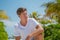 Handsome tanned young man wearing white t-shirt and black shorts sitting on the tropical beach at the island luxury resort