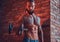 A handsome tattoed shirtless male with a stylish haircut and beard, doing exercise with a dumbbell, standing against a