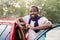 Handsome young African bearded man with smartphone near his red vehicle on outdoor parking, looking at camera and