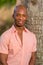 Handsome young black man posing by tree in the park. Man wearing a pink button shirt and smiling at the camera