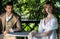 Handsome young couple in vintage clothing sit in gazebo. The female is looking at camera as her companion looks at her.