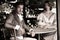 Handsome young couple in vintage clothing sit in gazebo with handsome gentleman looking at camera as companion looks on.