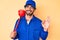 Handsome young man with curly hair and bear wearing plumber uniform holding toilet plunger doing ok sign with fingers, smiling