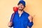 Handsome young man with curly hair and bear wearing plumber uniform holding toilet plunger pointing thumb up to the side smiling