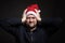 A handsome young man in a Santa Claus hat clasps his head with his hands and squints. Black background. Close-up