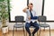 Handsome young man sitting at doctor waiting room with arm injury making fun of people with fingers on forehead doing loser