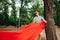 Handsome young man touches a red tourist hammock to a tree in the forest with a serious face