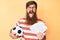 Handsome young red head man with long beard holding football ball and bunch of dollars sticking tongue out happy with funny