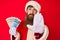 Handsome young red head man with long beard wearing santa claus costume holding canadian dollars with open hand doing stop sign