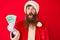 Handsome young red head man with long beard wearing santa claus costume holding israel shekels scared and amazed with open mouth