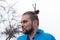 Hansome bearded man with hair bun with sticks and make up looking away in front of tree