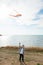 Happy active little adorable kid holding flying beautiful kite standing on sea beach during summer leisure vacation