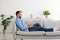 Happy adult european man with beard sitting on sofa and typing on laptop in living room interior, profile