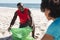 Happy african american couple cleaning sunny beach and collecting litter