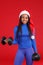 A happy African-American girl, dressed in Santa`s cap, holds heavy weights. On a red background.