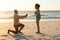 Happy african american man kneeling and proposing to girlfriend on beach at sundown